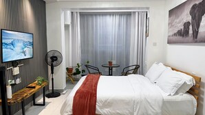 A modern hotel room with a bed, a TV, a fan, a small table with a plant, and a window with curtains.