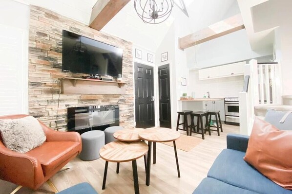 A modern living room with a stone wall, a flat-screen TV, a fireplace, a blue sofa, and a kitchen area in the background. - Rustic Loft at Blue (Blue Mountain)