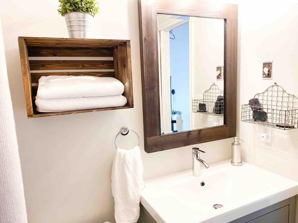 Un baño con un estante de madera con toallas, un espejo grande, un lavamanos blanco y una jaula de pájaros en la pared.