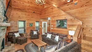 A cabin interior with wooden walls, a chandelier, a fireplace, and a sofa set.