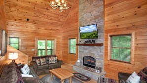 A cozy cabin interior with a stone fireplace, wooden walls, a television mounted above it, and a comfortable seating area with a sofa and armchairs.