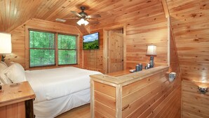 A wooden cabin bedroom with a bed, a desk, a lamp, a ceiling fan, and a TV on the wall.