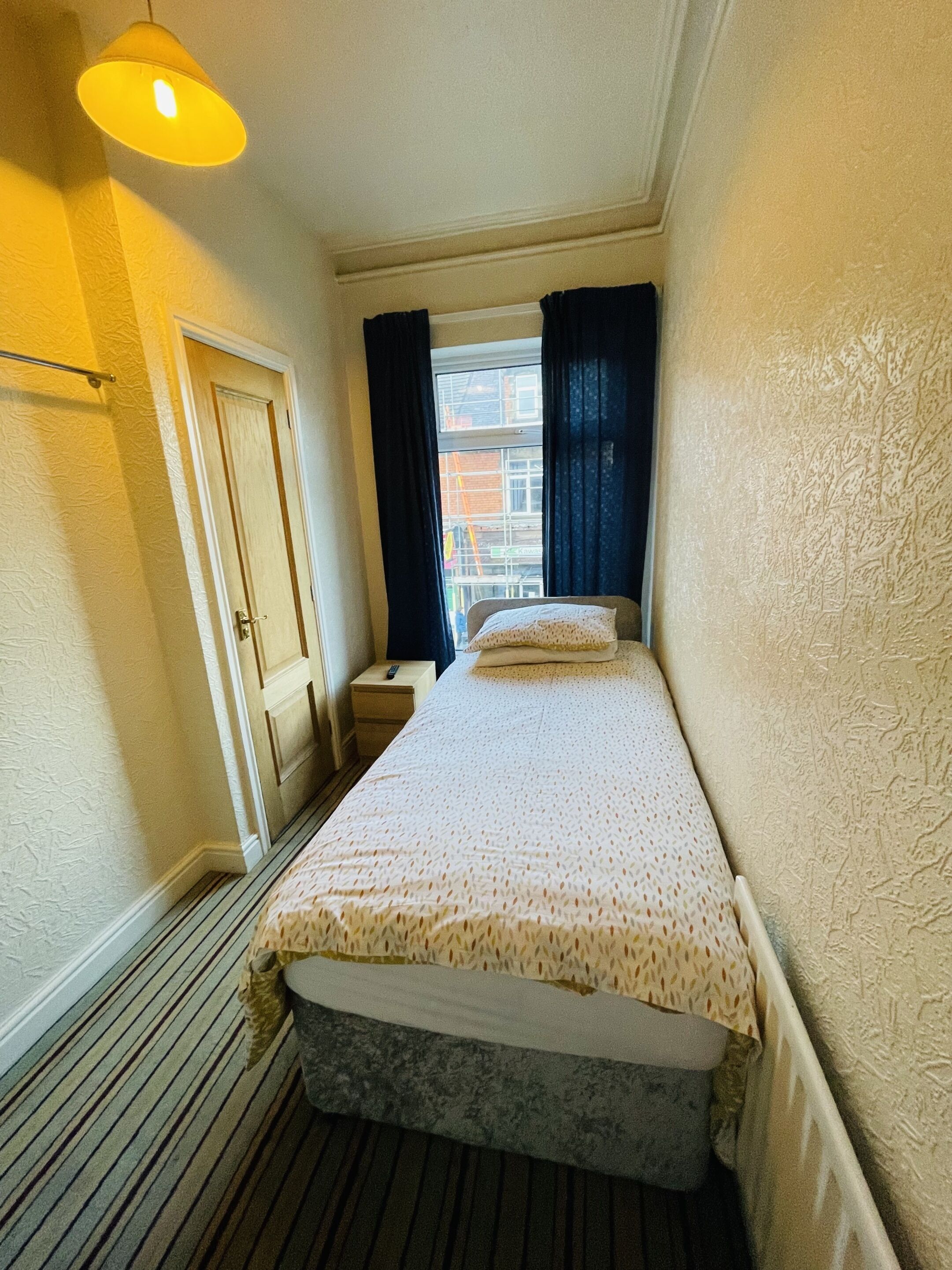 A small, narrow hotel room with a single bed, a bedside table, a nightstand, a window with curtains, and a radiator.
