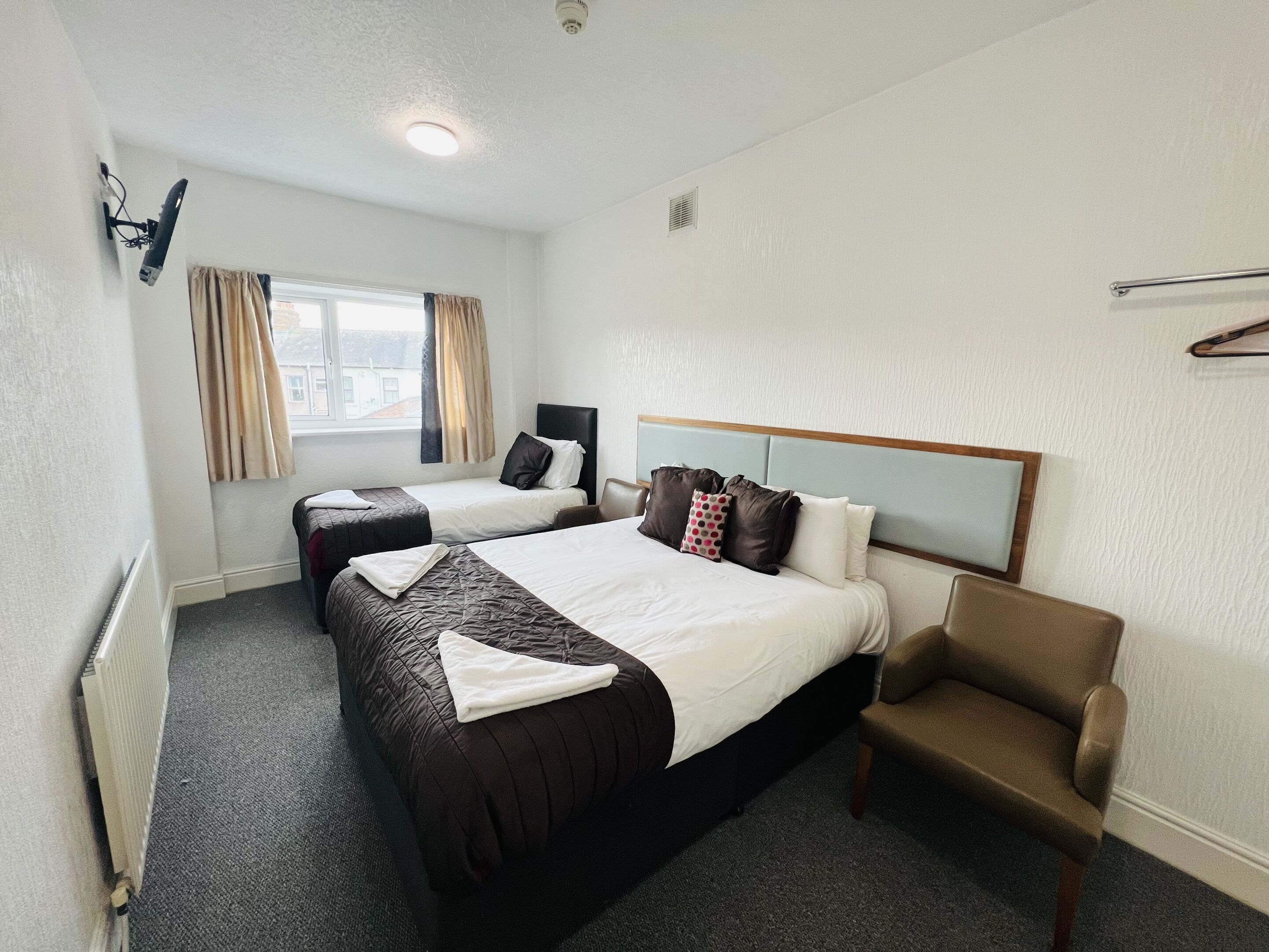 A hotel room with two beds, a chair, a window with curtains, a radiator, and a ceiling light.