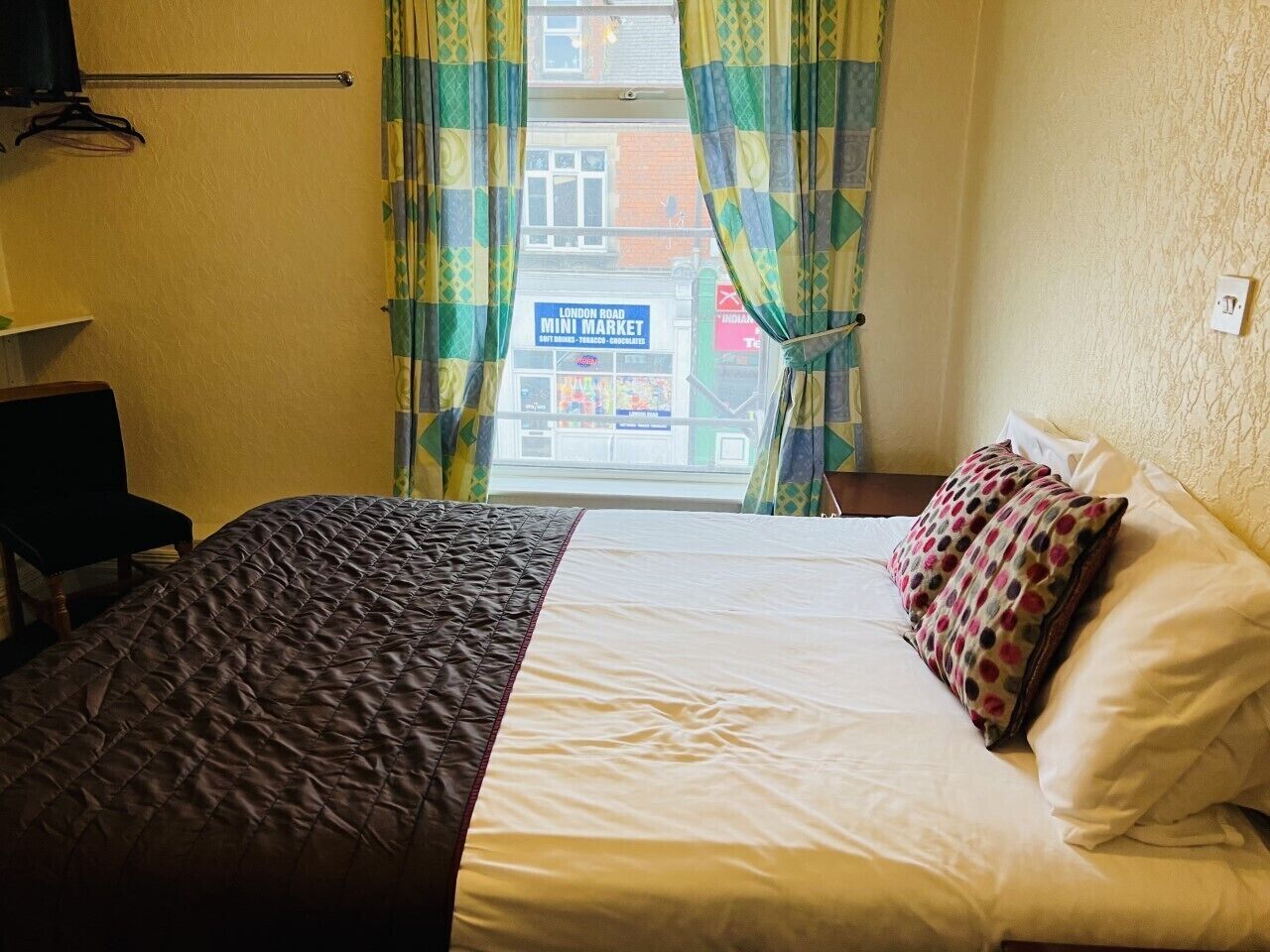 A bedroom with a bed, a chair, a TV, and a view of a mini market through the window.