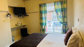 A hotel room with a bed, a TV mounted on the wall, a chair, a small shelf with toiletries, and a window with curtains.