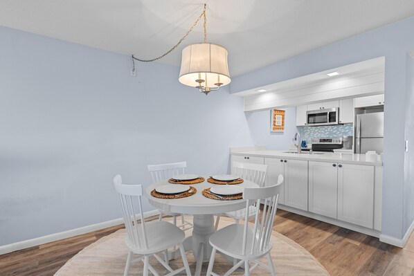 A modern kitchen with white cabinets, a microwave, and a refrigerator. There is a dining table with four chairs and a hanging light fixture.