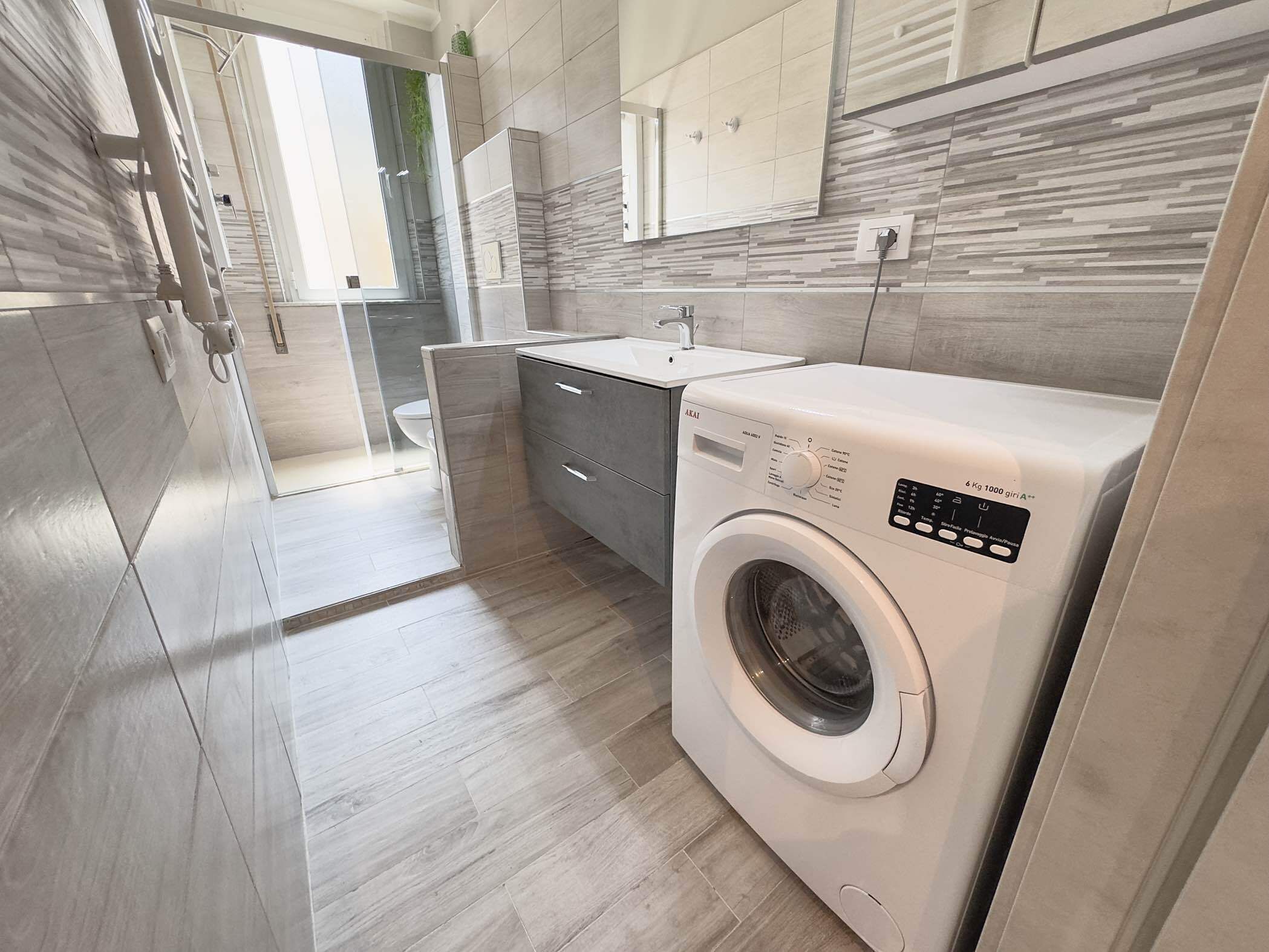 A modern laundry room with a front-loading washing machine, a small sink, and a glass shower enclosure.