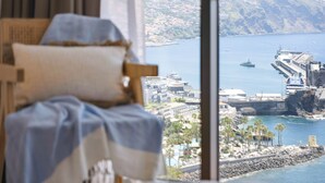 A room with a large window overlooking a coastal area, including a harbor, buildings, and a rocky shoreline. - Infinity View by Madeira Sun Travel (Funchal)