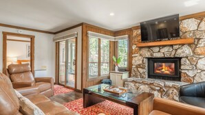 A living room with leather sofas, a glass coffee table, a stone fireplace, and a TV mounted above it.