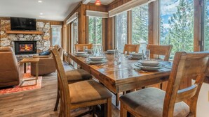 A dining area with a wooden table set for six, surrounded by chairs. A stone fireplace with a TV above it is visible, and large windows offer a view of trees and mountains.