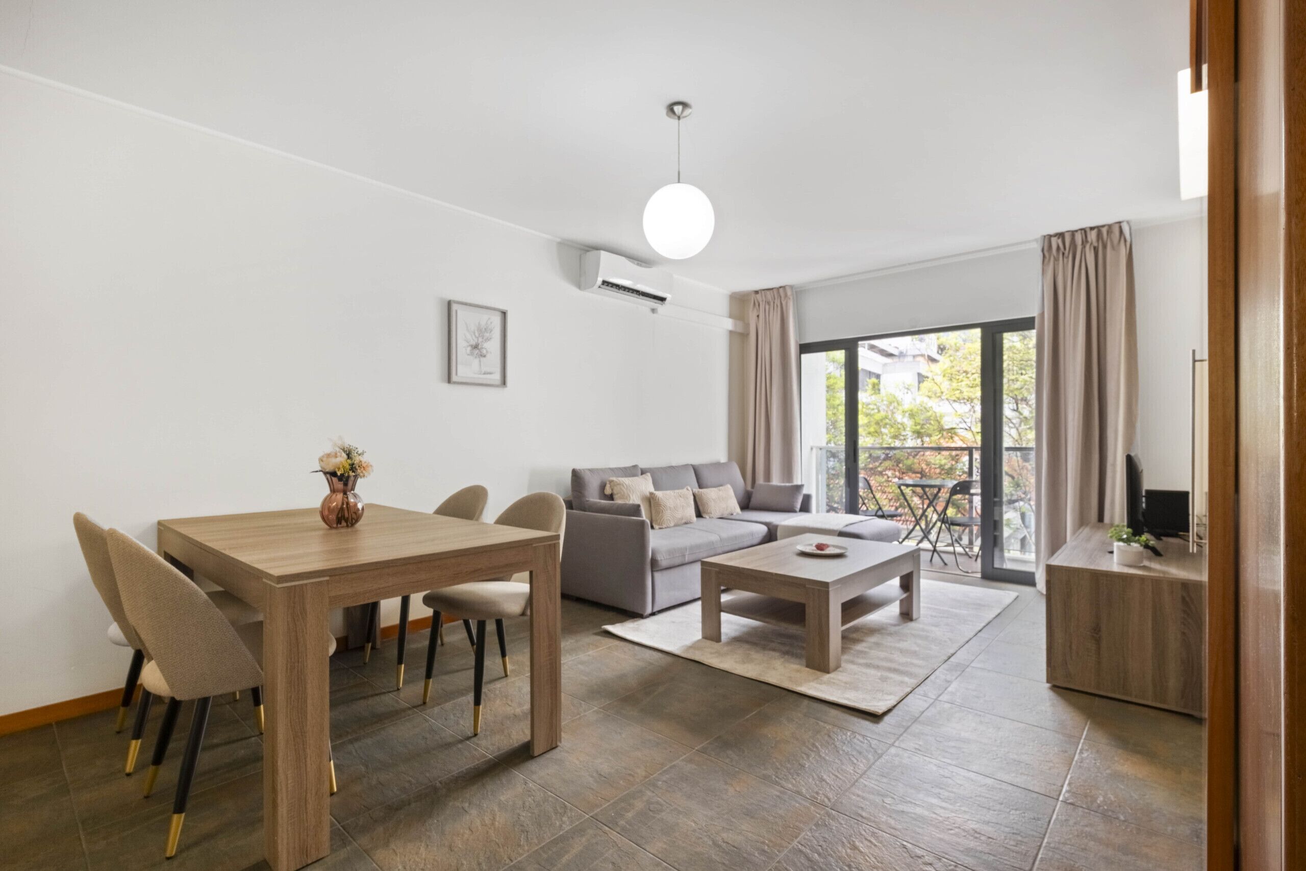 Een moderne woonkamer met een eettafel, bank en salontafel. Er is een groot raam met gordijnen, een televisie op een houten standaard en een airconditioning aan de muur.