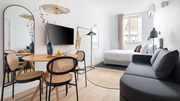 A modern living room with a grey sofa, a dining table with wicker chairs, a flat-screen TV, and a large mirror.