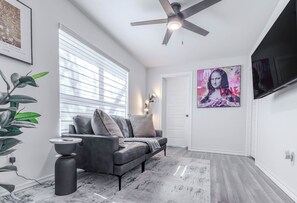 A modern living room with a grey sofa, a large window with blinds, a ceiling fan, and a painting on the wall.