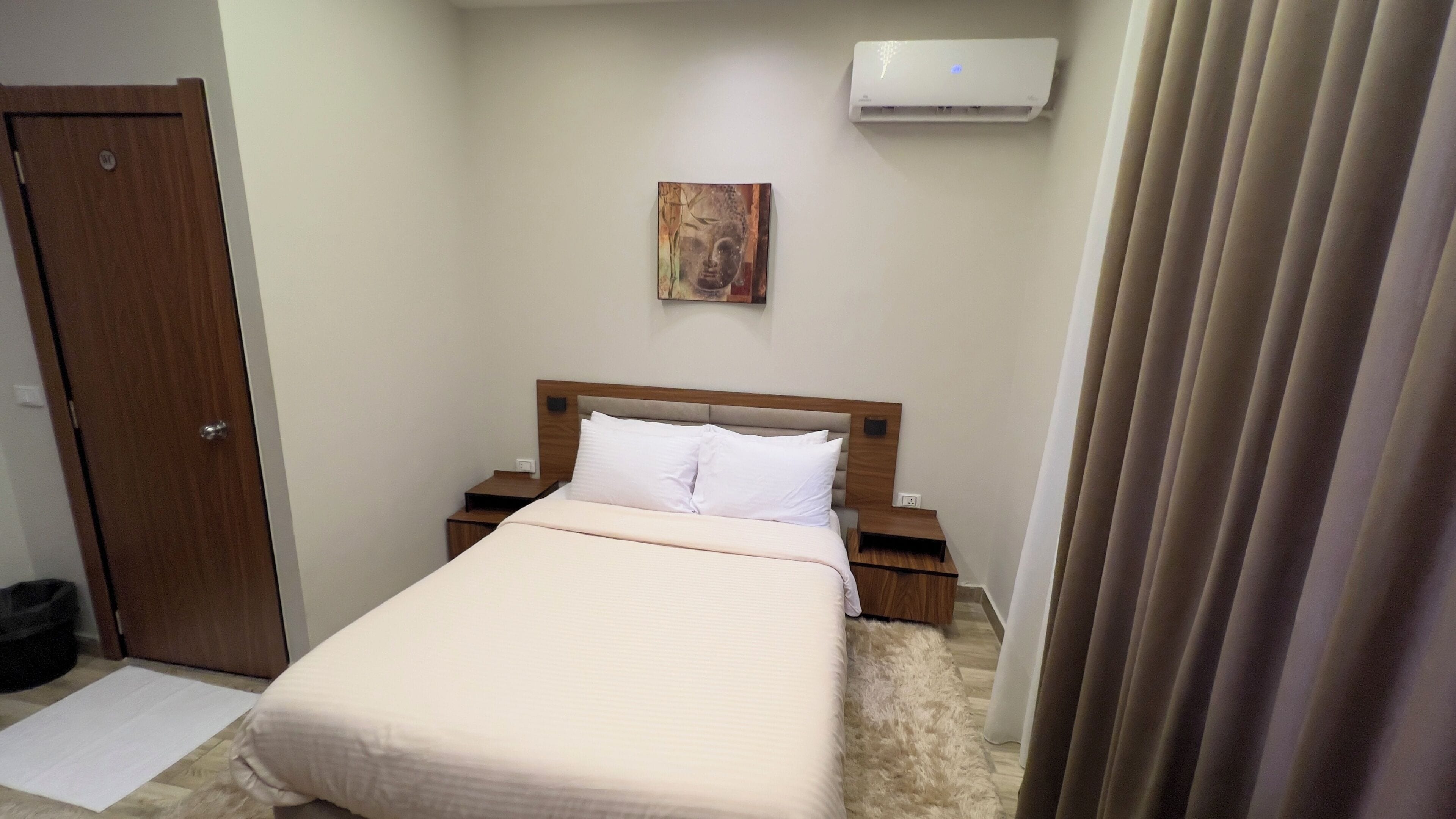 A hotel room with a bed, a nightstand, a wall-mounted air conditioner, and a door.