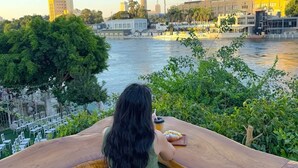 A person seated at a wooden table overlooking a river with a cityscape in the background. - Italia house hotel (Cairo)