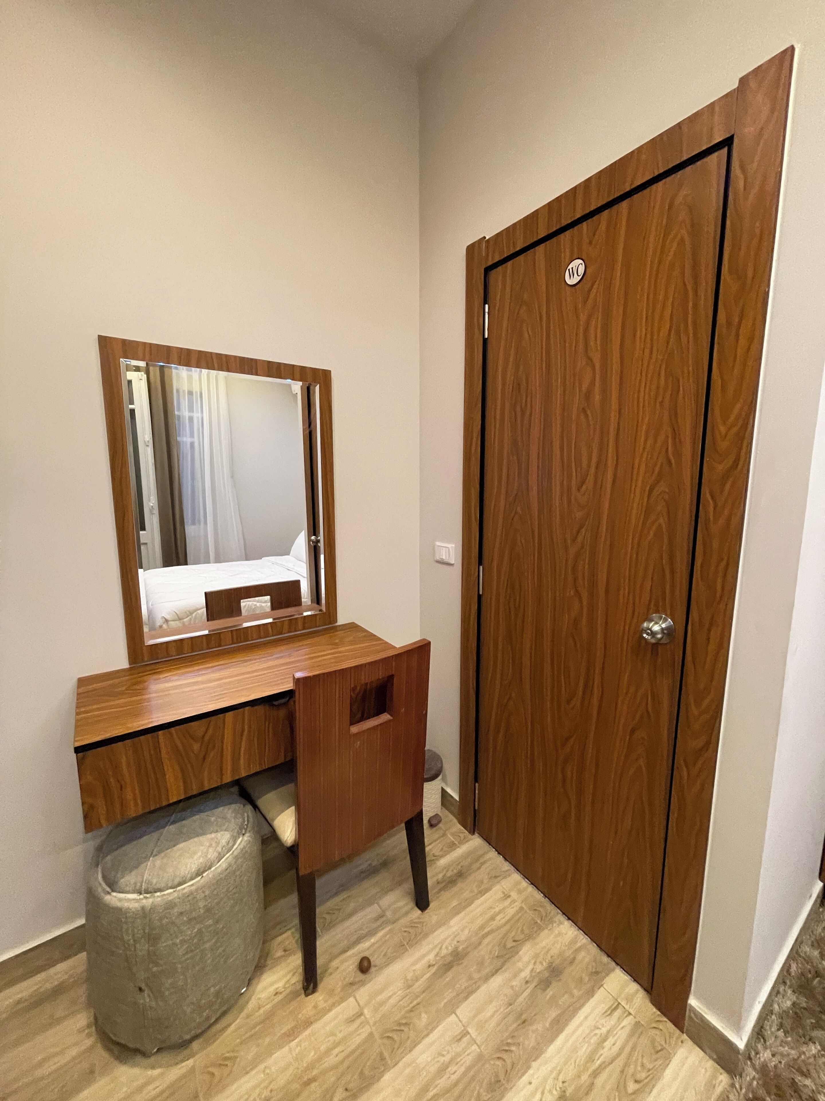 A compact room with a wooden desk, a mirror, a small trash bin, and a wooden door.