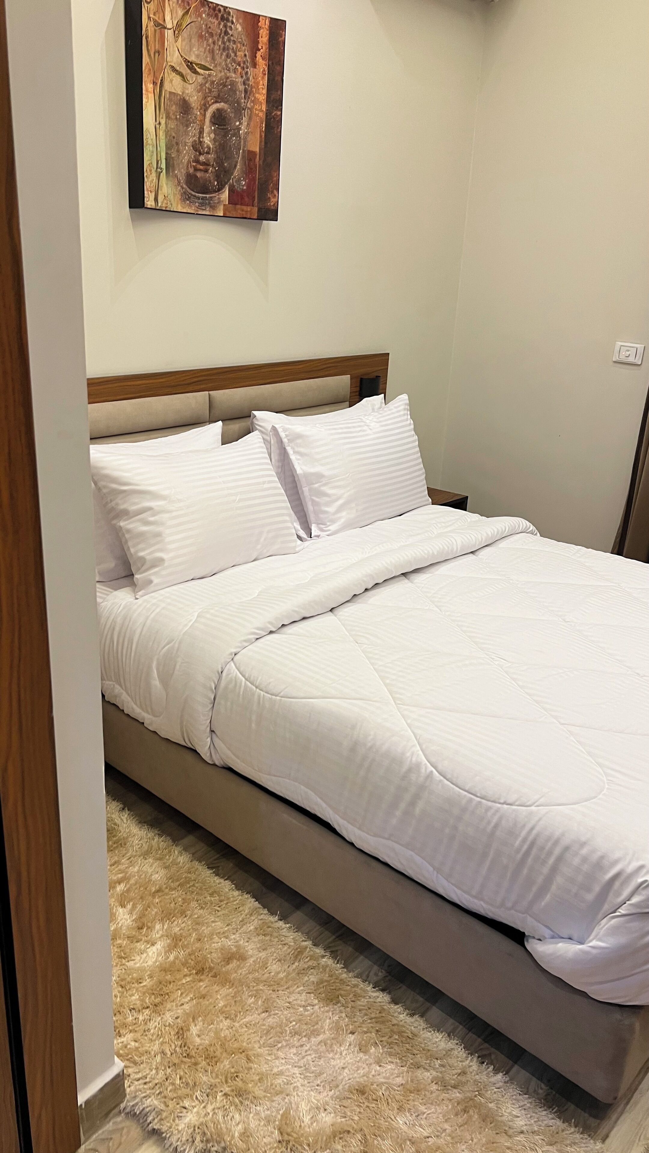 A neatly made bed with white linens, a wooden headboard, and a fluffy beige rug.