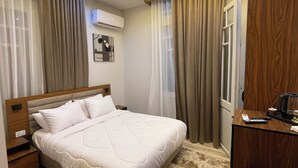 Quarto moderno com uma cama grande, uma mesa de trabalho e uma cadeira. Há uma janela com cortinas, um aparelho de ar condicionado e uma obra de arte na parede.