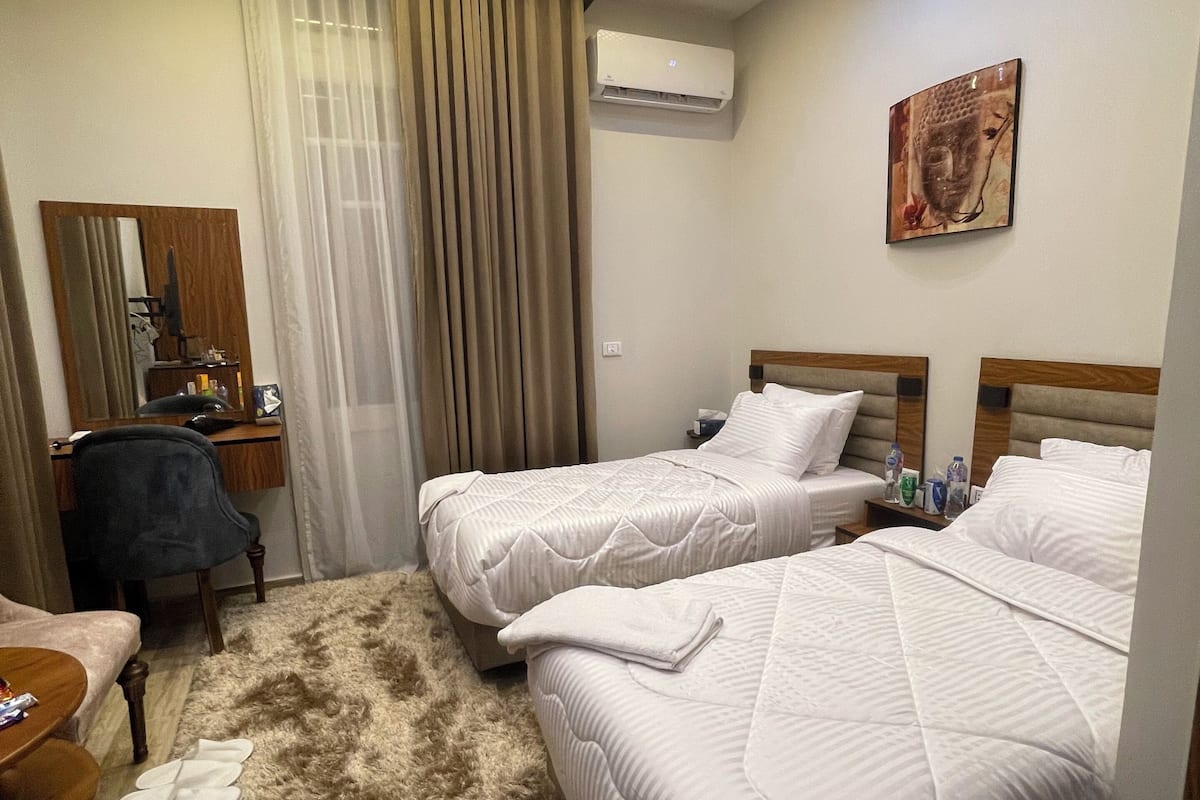 Ein Hotelzimmer mit zwei Betten, einem Schreibtisch, einem Stuhl, einem Spiegel, einem Fenster mit Vorhängen und einer Klimaanlage.