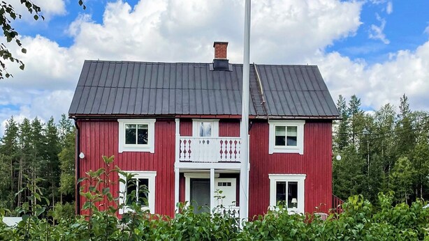 A red two-story house with a grey roof, surrounded by greenery.