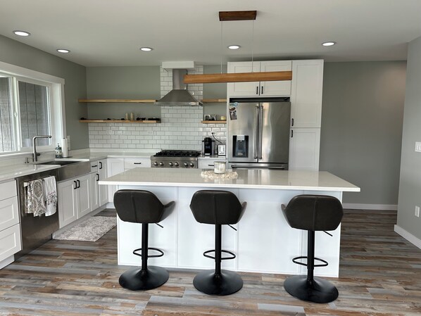 A modern kitchen with white cabinets, a stainless steel refrigerator, a gas stove, and a bar area with three stools. - Water and mountain view’s in charming Juneau (Juneau)