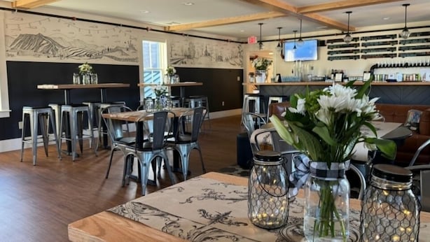 Un restaurant avec un plafond haut, des poutres apparentes en bois et un coin bar équipé d’un téléviseur. Les tables sont disposées avec des chaises et décorées de fleurs et de lanternes.
