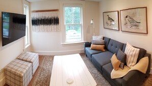 Un salon moderne avec un canapé gris, une table basse blanche et une grande fenêtre donnant sur des arbres. On y trouve deux poufs, un téléviseur fixé au mur et, sur le mur, des illustrations d’oiseaux encadrées.