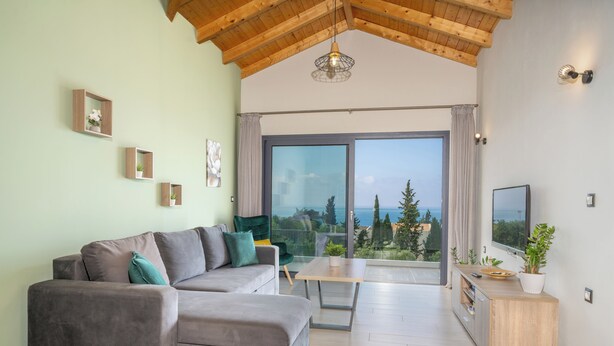 Ein modernes Wohnzimmer mit einem grauen Ecksofa, einem Holztisch und einer großen Glasschiebetür, die auf einen Balkon mit Blick auf Bäume und das Meer führt.