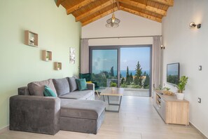 Un salon moderne avec un canapé d’angle gris, une table basse en bois et une grande porte-fenêtre coulissante donnant sur un balcon avec vue sur les arbres et la mer.