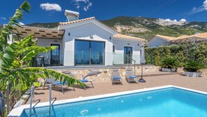 Ein modernes Haus mit Swimmingpool, einer Terrasse mit Liegestühlen und einem Berg im Hintergrund.