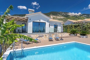 Una casa moderna con piscina, un patio con sillas de descanso y un telón de fondo de montañas.