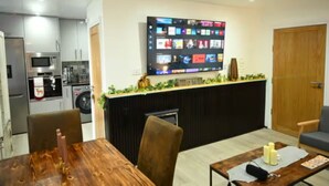 A modern kitchen with a built-in microwave, oven, and washing machine. There is a dining area with a wooden table and chairs. A flat-screen TV is mounted on the wall above the counter. - Watermead Retreat Bungalow (Leicester)