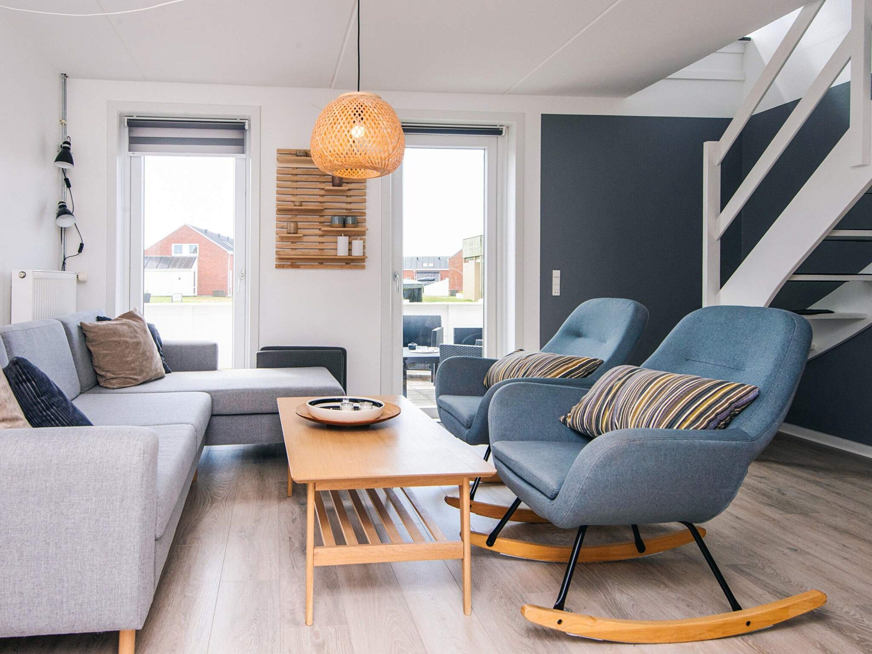 Ein modernes Wohnzimmer mit einem grauen Sofa, einem blauen Schaukelstuhl, einem Holztisch und einem großen Fenster mit Blick auf Gebäude.