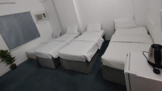 A hotel room with two single beds, a small desk with a kettle, a potted plant, and a window with blinds. - Al Awali Economic Apartments (Madinah)