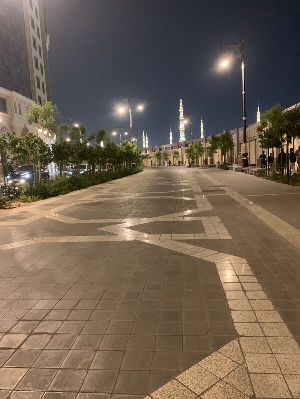 A spacious plaza with tiled flooring, street lamps, and a mosque with illuminated minarets in the background.