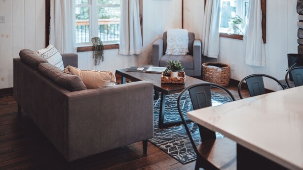 A cozy living room with a sofa, a dining table, and a chair. There is a window with curtains, a plant, and a basket on the table. - Chapter's End Luxury Retreat (Logan)