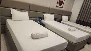 Ein Hotelzimmer mit drei Betten, jeweils mit weißen Laken und Kissen. Auf den Betten liegen Handtücher, auf dem Nachttisch eine Wasserflasche und an der Wand ein gerahmtes Bild.