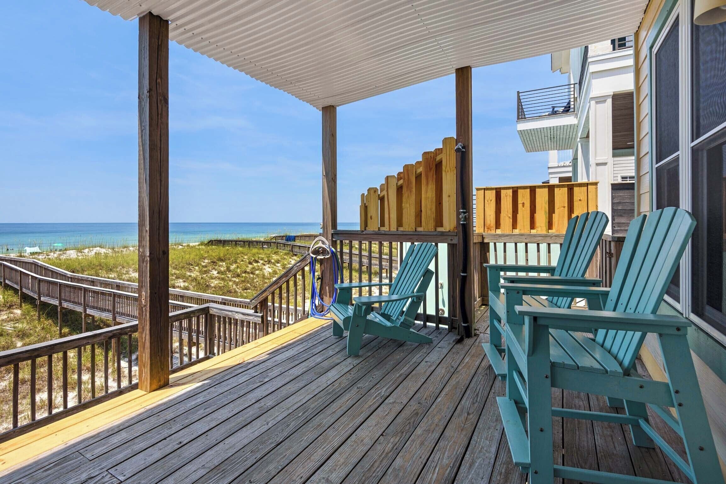 A wooden deck with a railing overlooking a beach, featuring two green Adirondack chairs and a wooden fence.