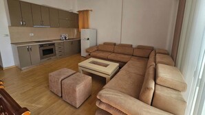 A modern living room with a large sectional sofa, a small coffee table, and a kitchen area with cabinets and appliances. - Albania Dream Saranda Apartments (Sarandë)