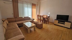 A modern living room with a sectional sofa, a coffee table, a dining table with chairs, and a TV on a cabinet. - Albania Dream Saranda Apartments (Sarandë)