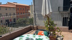 Eine Dachterrasse mit Tisch, bedeckt mit einer Tischdecke mit Blätermotiv, zwei Stühlen, einem Blumentopf und einem weißen Sonnenschirm.