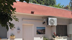 Ein Gebäude mit weißer Fassade, rotem Dach und einem Balkon mit Metallgeländer. Im Erdgeschoss stehen Blumentöpfe und ein Wandgerät zur Klimatisierung.