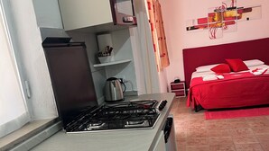 Eine kompakte Küche mit Gasherd, Spüle und kleinem Kühlschrank. Im angrenzenden Raum ist ein Bett mit roter Bettwäsche zu sehen.