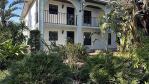 Un edificio de dos pisos con un balcón en el segundo nivel, rodeado de exuberante vegetación tropical.