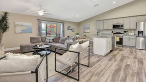Un vaste salon comprenant un ensemble de canapés, une table basse et un coin repas avec une table et des chaises. La cuisine est équipée d’appareils électroménagers en acier inoxydable et d’un îlot central.