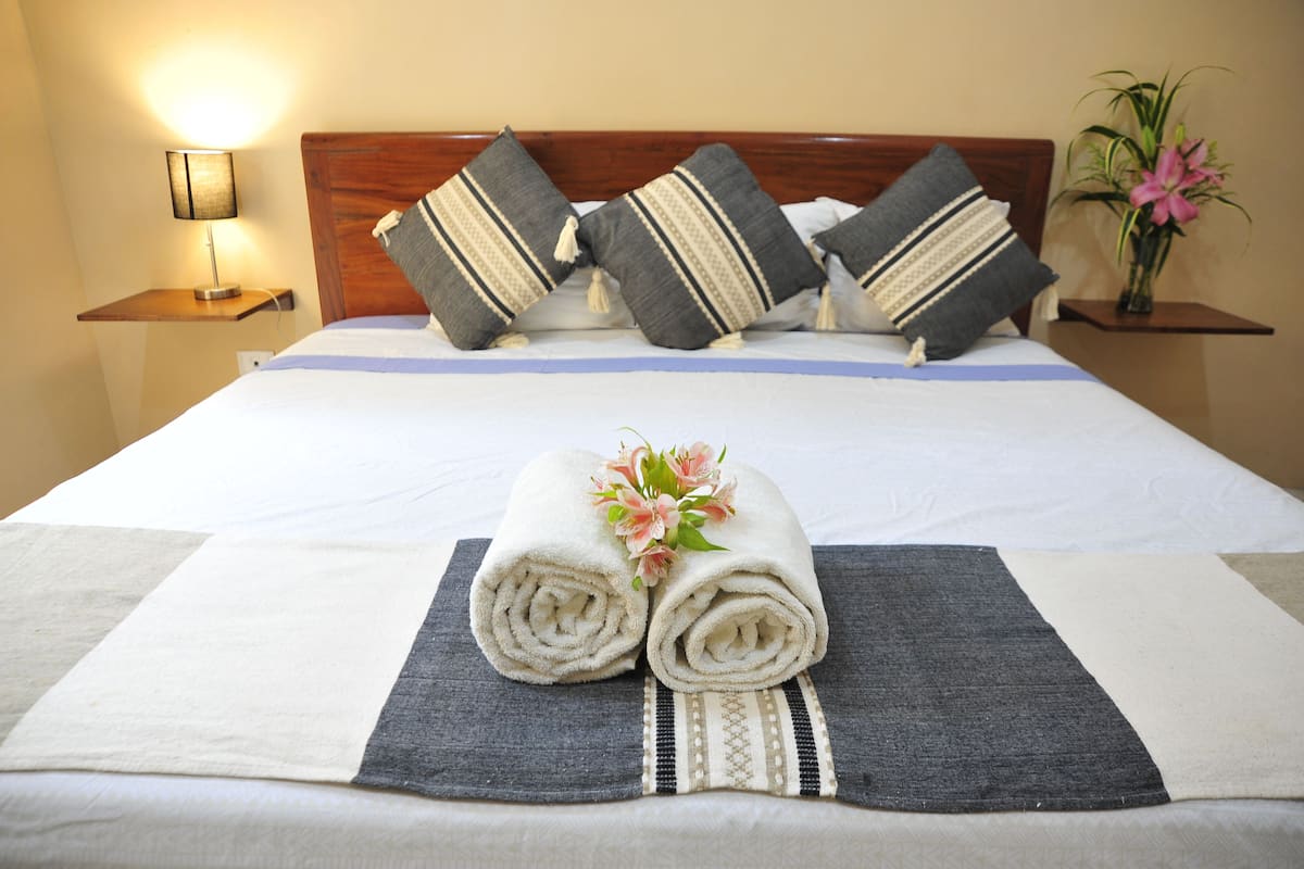 A neatly made bed with a rolled towel and a flower, a bedside lamp, and a vase with flowers on a nightstand.