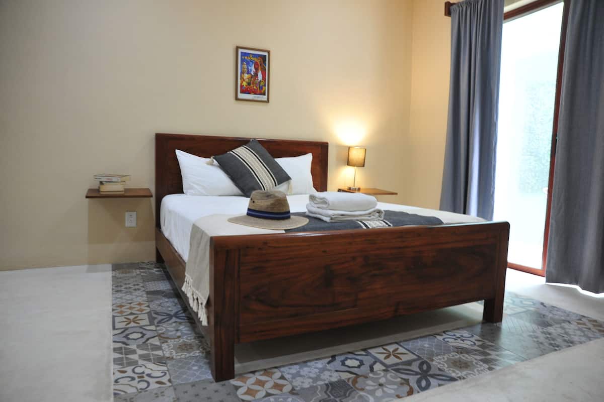 A neatly made bed with a wooden headboard, a bedside lamp, and a small table with books and a hat.
