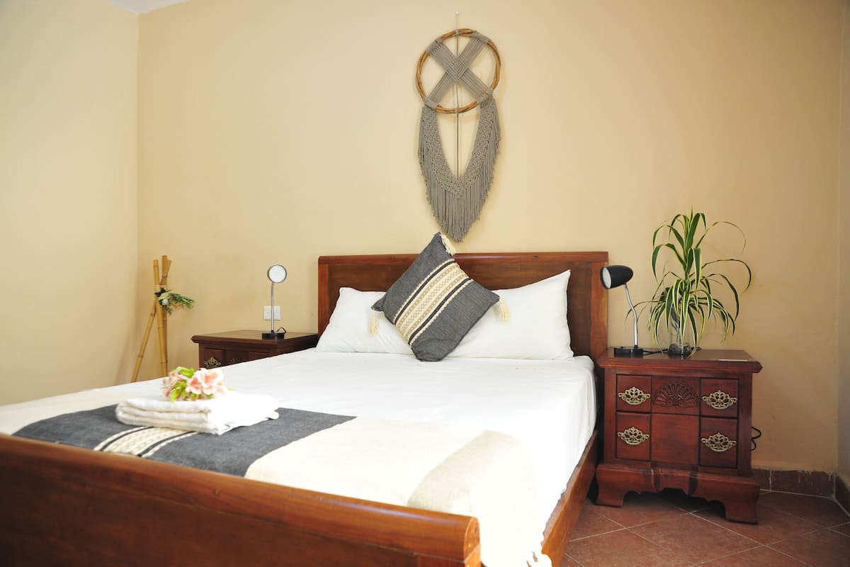 A neatly made bed with a wooden headboard, a bedside table with a lamp, and a wall-mounted macrame decoration.