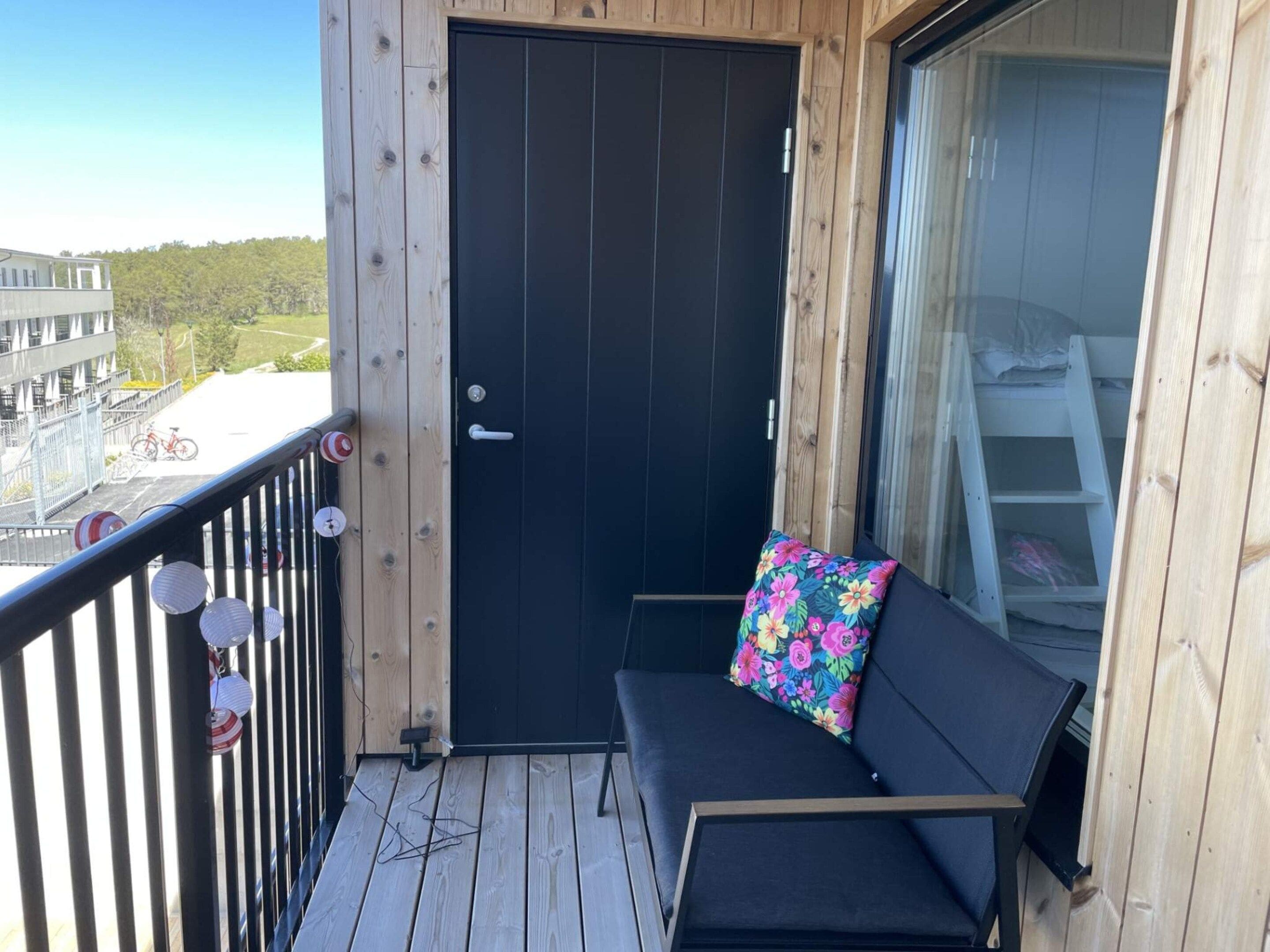 A balcony with a black door, a bench, and a view of a building and trees.